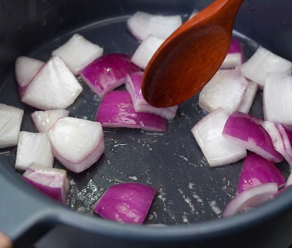 Trozos de cebolla roja picada siendo removidos con una cuchara de madera en una olla oscura a fuego medio.