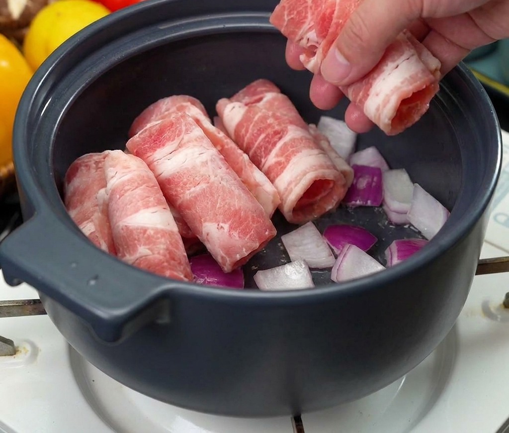 Una mano colocando varios rollos de carne de res cruda finamente rebanada en una olla sobre una base de cebollas sofritas.