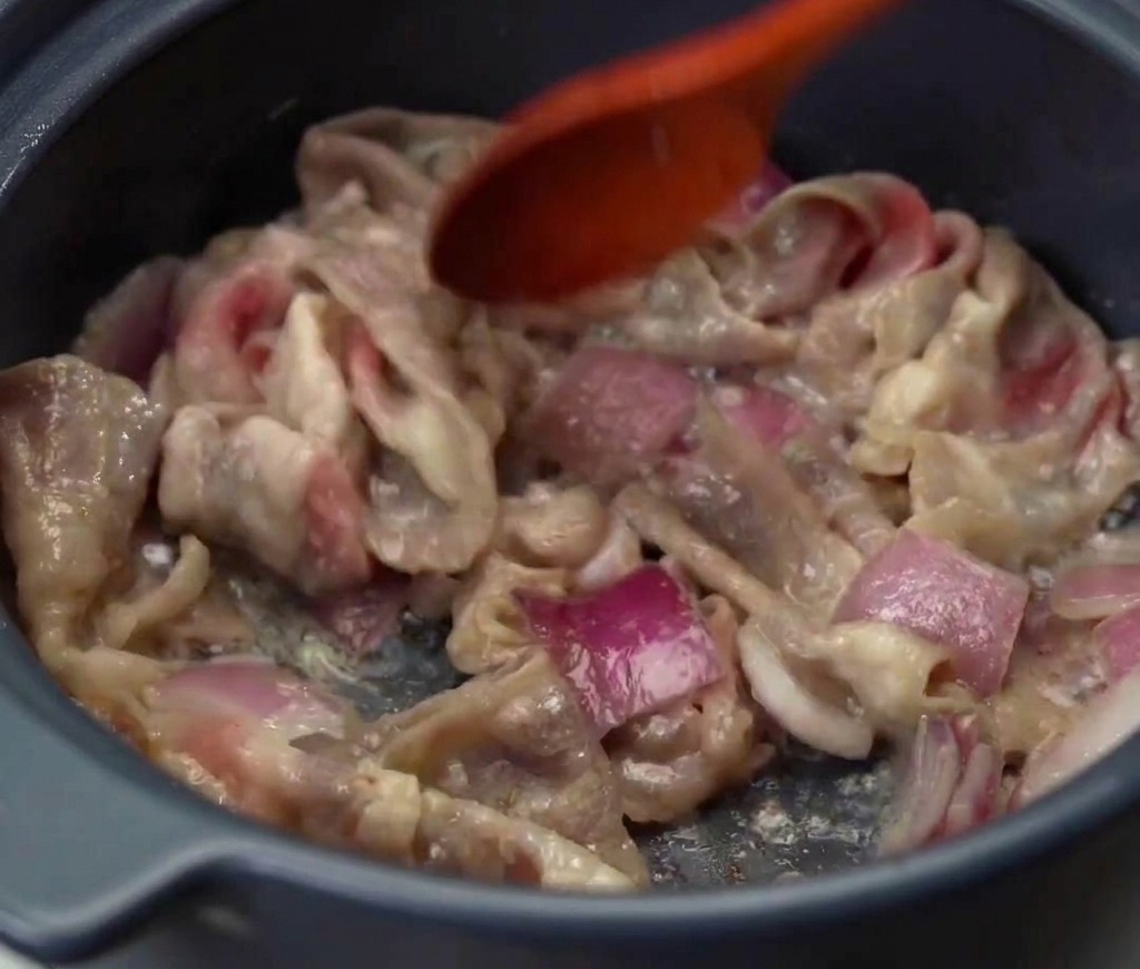 Carne de res en láminas y cebollas rojas siendo salteadas juntas en una olla hasta que la carne esté cocida y dorada.
