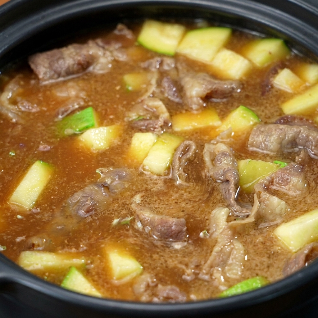 La pasta de soja mezclándose y disolviéndose en la olla, transformando el líquido en un caldo marrón opaco con trozos visibles de carne y calabacín.