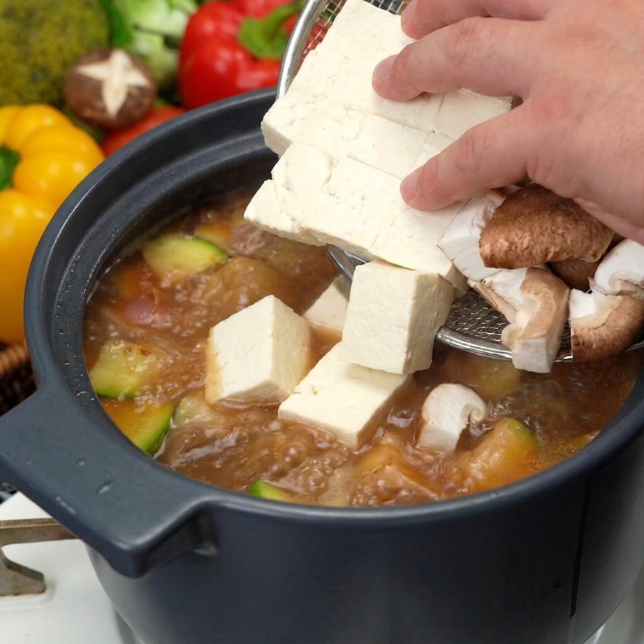 Una mano usando un colador para añadir cubos de tofu blanco fresco y láminas de hongos shiitake marrones a una olla de sopa de pasta de soja hirviendo.