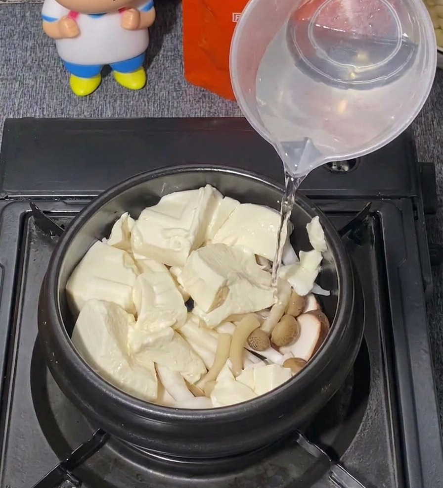 Agua limpia vertiéndose desde una taza medidora de plástico en una olla negra llena de tofu y hongos.
