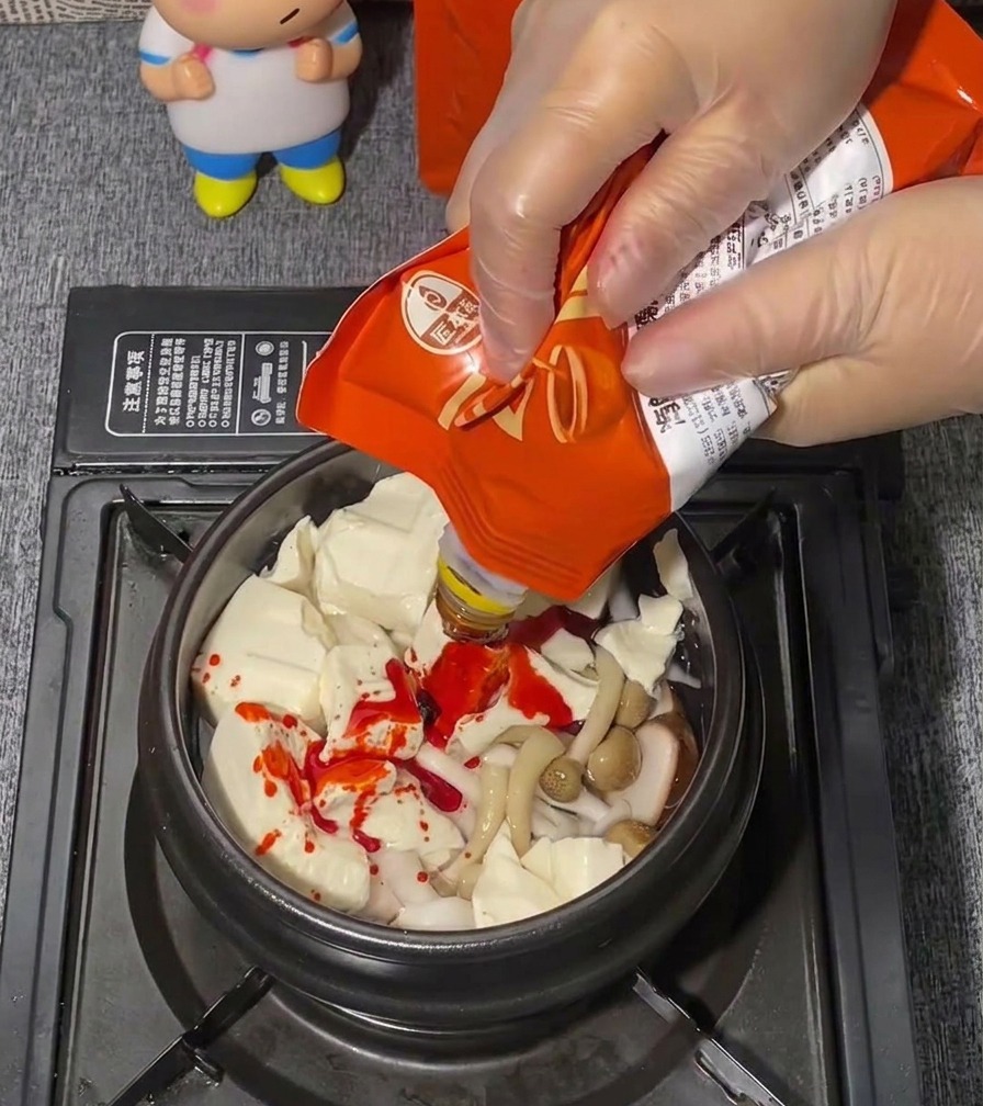 Una mano con un guante de plástico transparente exprimiendo una pasta de sazonador rojo brillante desde una bolsa sobre una cama de tofu blando.