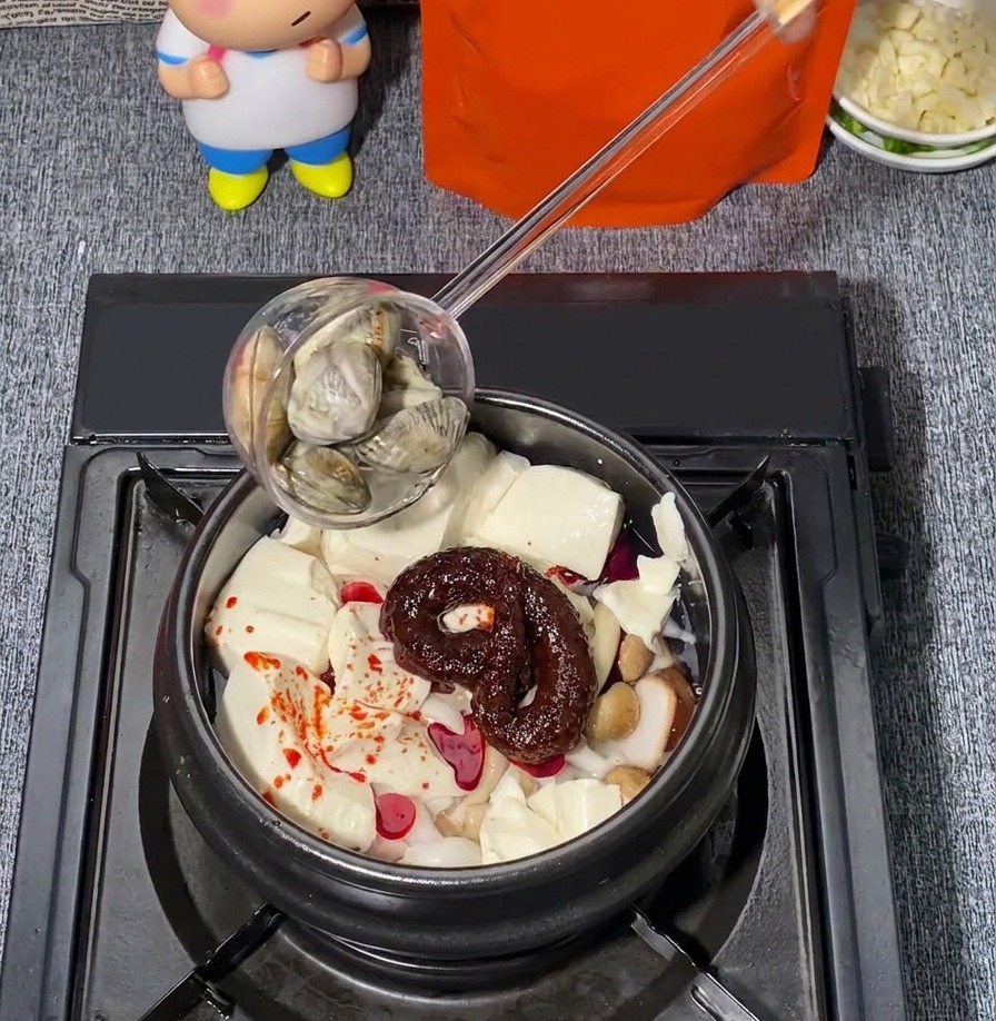 Almejas frescas añadiéndose con un cucharón en una olla de piedra que contiene tofu blando, hongos y sazonador de chile rojo.