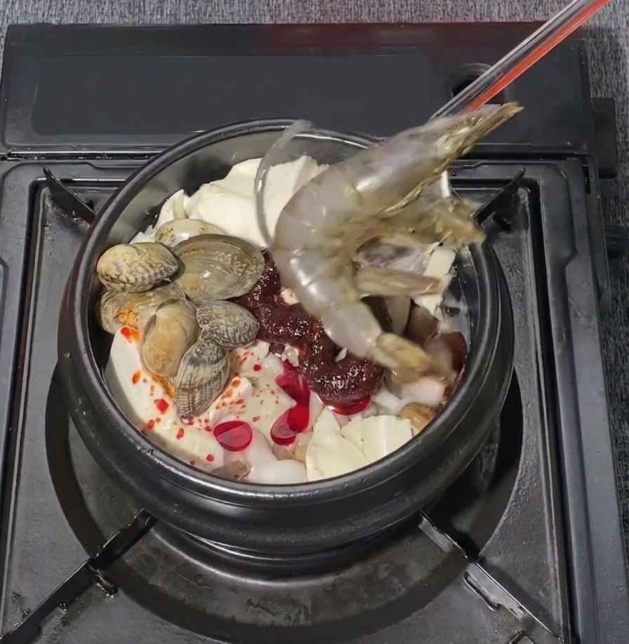 Dos langostinos grandes crudos colocándose con cuidado en una olla hirviente de estofado coreano de tofu blando con mariscos.
