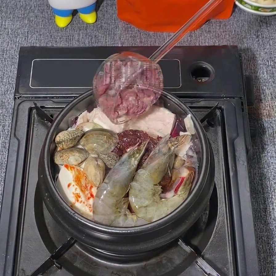 Finas láminas de ternera cruda añadiéndose desde un cucharón a una olla de estofado coreano que ya contiene langostinos y almejas.