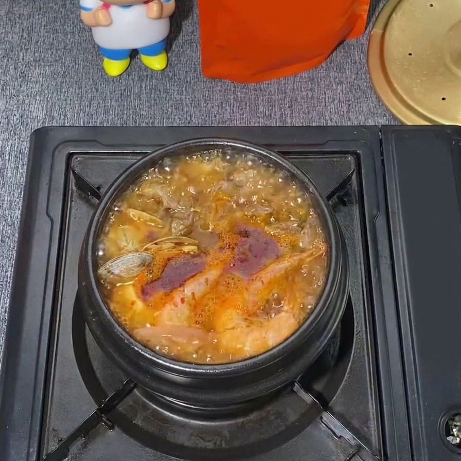 Una olla hirviendo a borbotones de estofado coreano de tofu blando con mariscos, con un caldo rojo brillante, almejas visibles y vapor subiendo.
