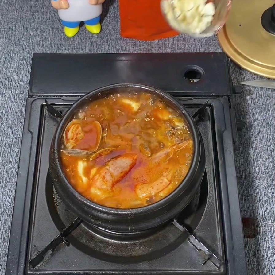 Un cucharón transparente dejando caer una cucharada de ajo blanco picado en el centro de un estofado coreano picante y rojo que está hirviendo.