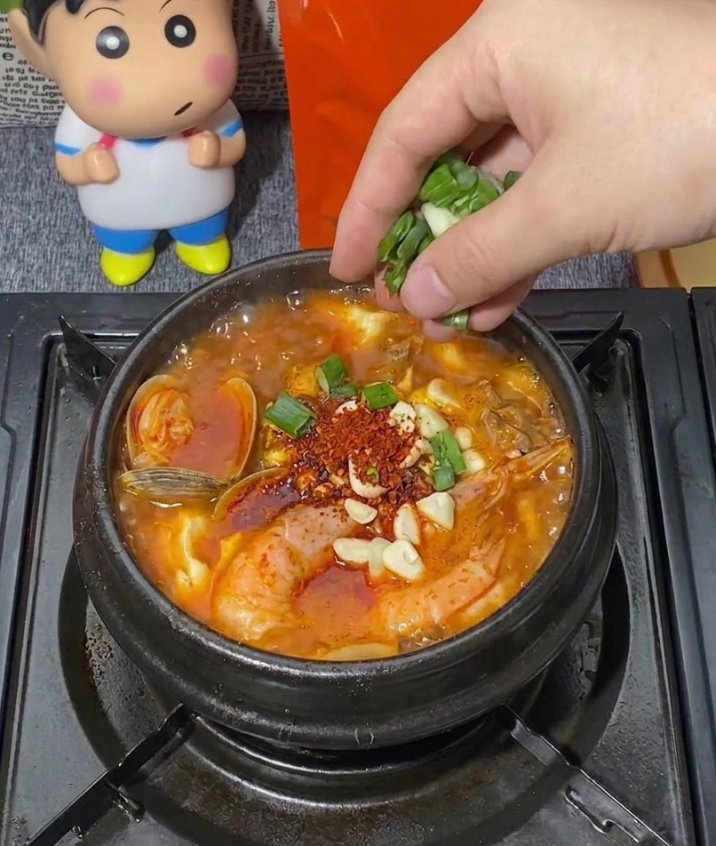Una mano espolvoreando un puñado de cebolleta verde picada en una olla de piedra negra hirviente con estofado de mariscos coreano lleno de camarones, almejas y ajo picado.