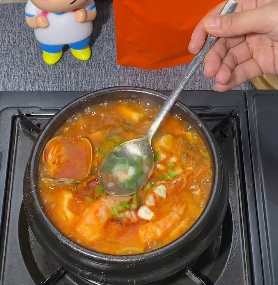 Una cuchara de metal removiendo cebolleta verde picada en una olla hirviente de estofado rojo de tofu blando con mariscos.