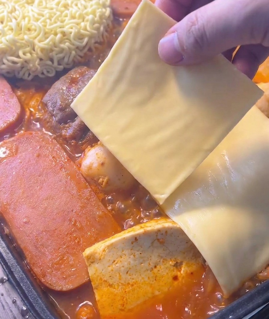 Una mano colocando una rebanada de queso amarillo sobre fideos instantáneos en un guiso rojo burbujeante