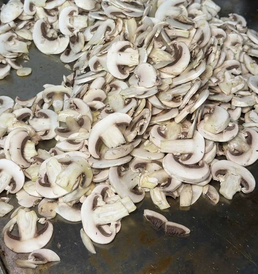 Champiñones laminados repartidos sobre una plancha metálica caliente desprendiendo vapor al empezar a cocinarse.