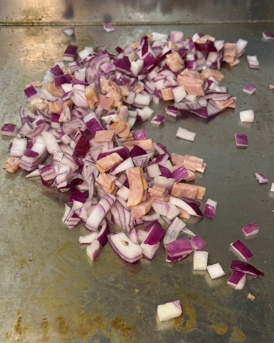 Mezcla de cebolla morada picada y trozos pequeños de bacon salteándose juntos sobre la superficie de una plancha metálica.