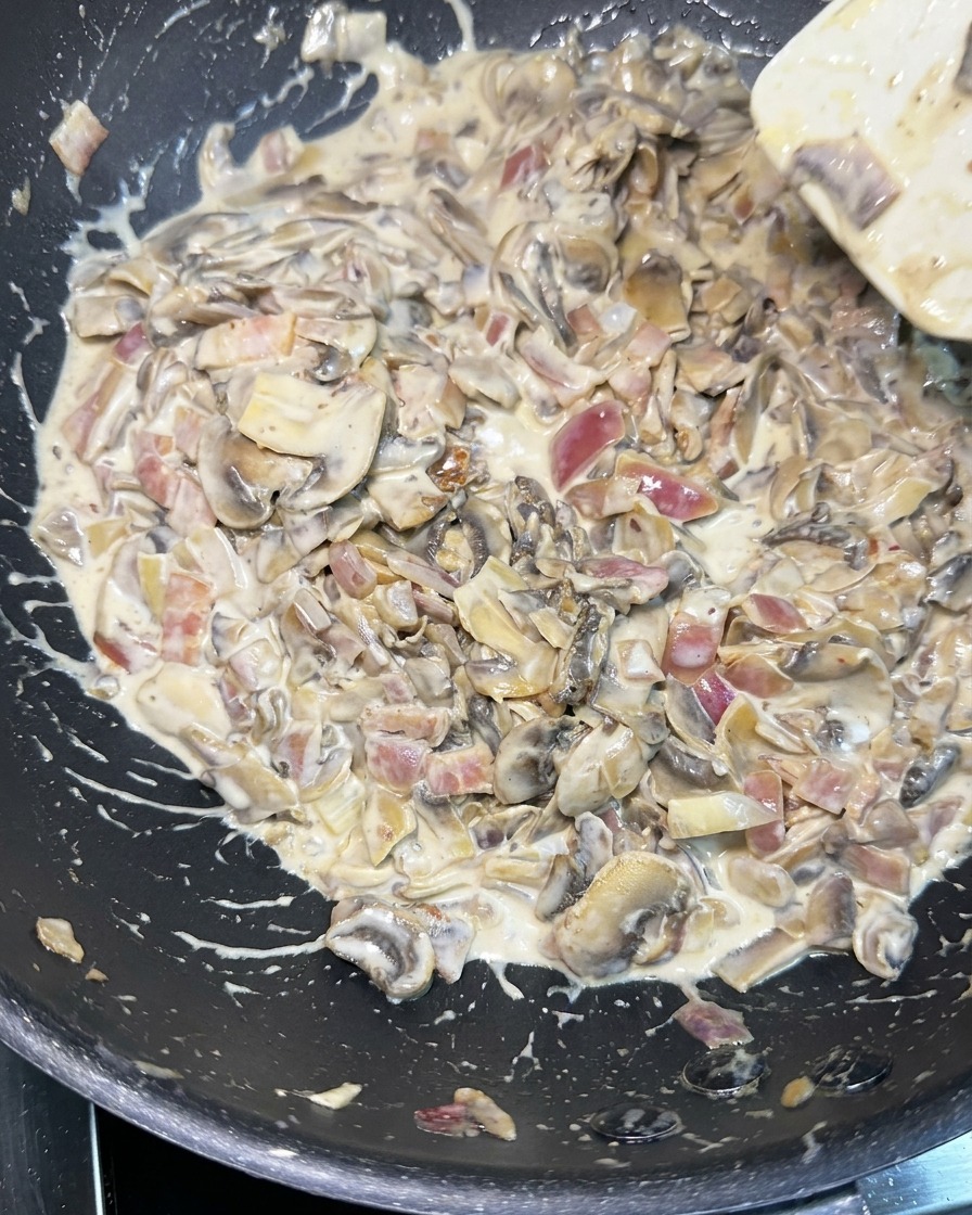 Una espátula blanca removiendo una salsa espesa y cremosa con champiñones laminados, cebolla picada y trozos de bacon en una sartén negra.