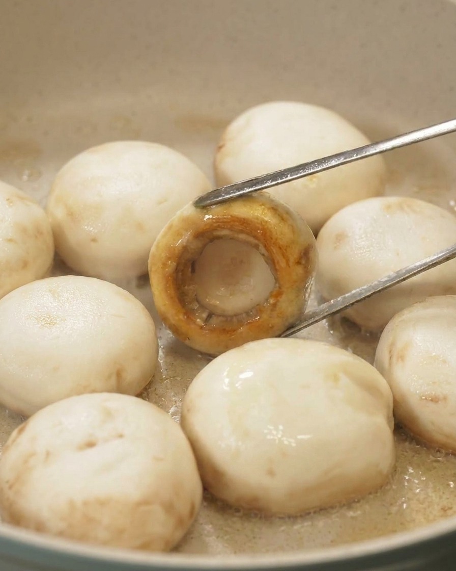 Pinzas volteando un champiñón en la sartén mostrando una superficie perfectamente caramelizada y dorada