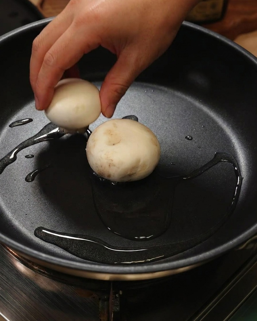 Una mano colocando un champiñón relleno boca abajo con la pasta de camarón hacia la sartén con aceite