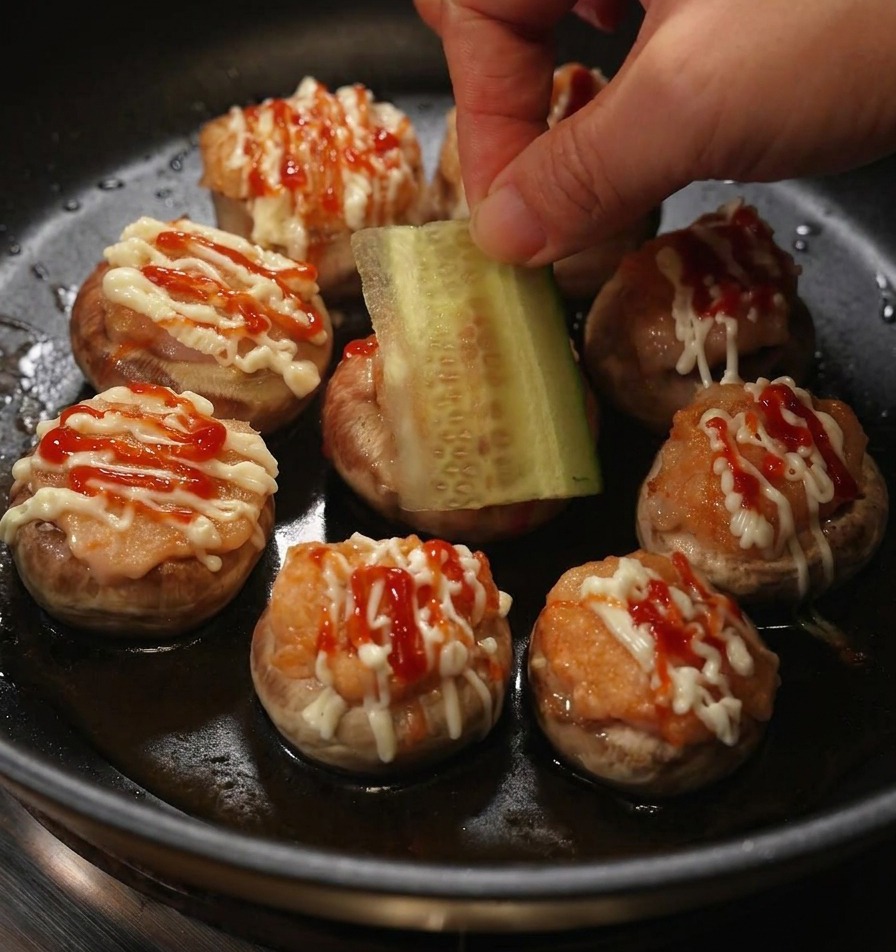 Una mano colocando una rodaja fina de pepino fresco sobre una mini hamburguesa de champiñón con mayonesa y kétchup