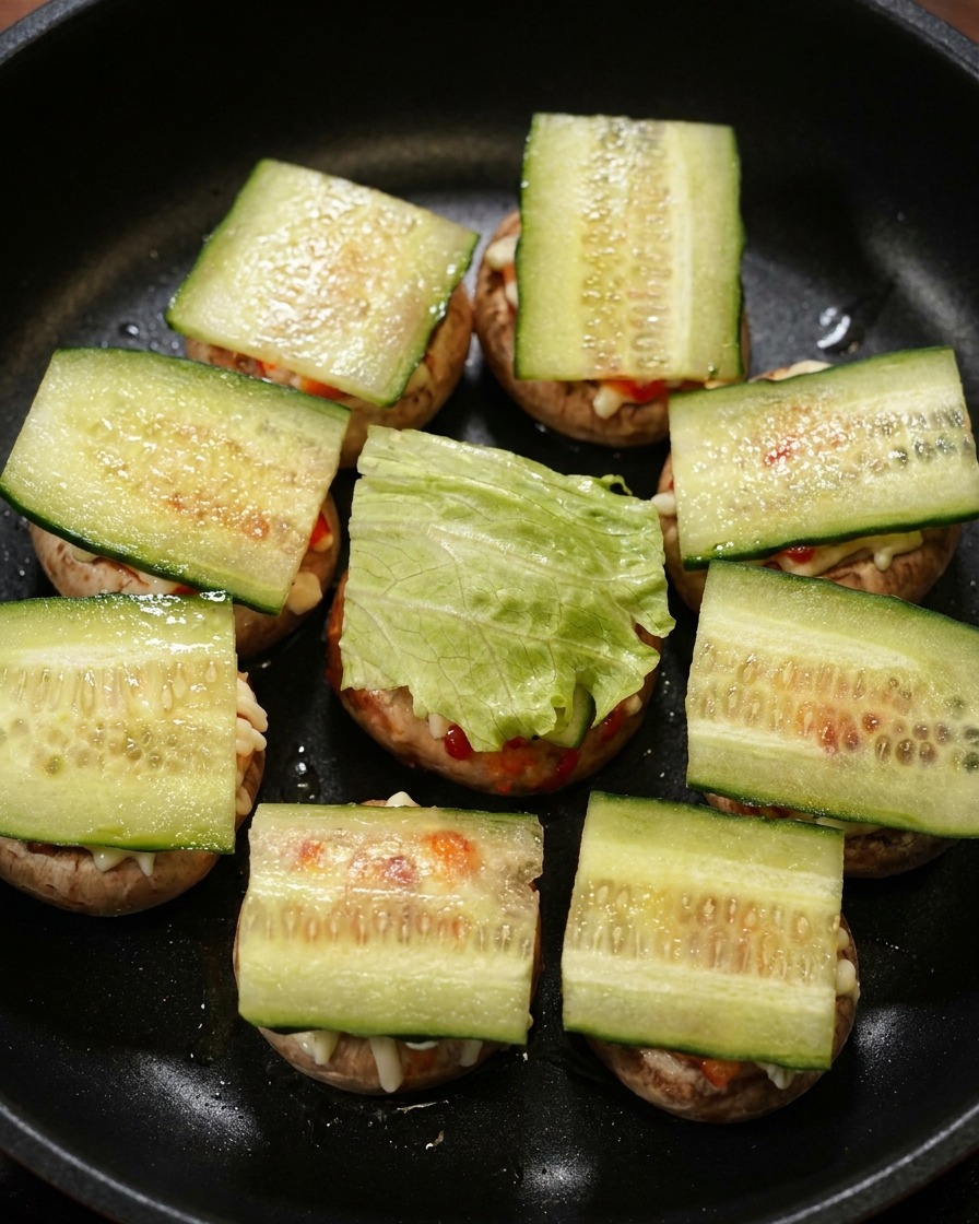 Rodajas de pepino y hojas de lechuga verde colocadas sobre las mini hamburguesas de champiñón en una sartén negra