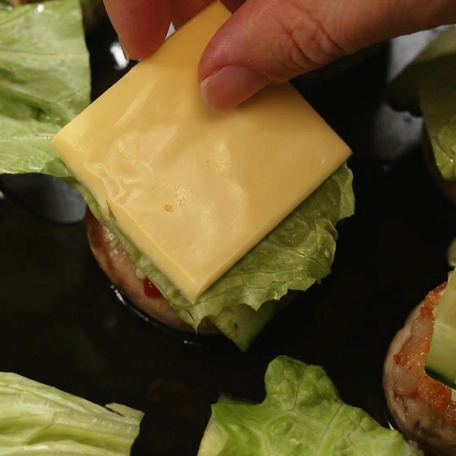 Una mano colocando un cuadrado de queso amarillo sobre la lechuga y el pepino dentro de la hamburguesa de champiñón