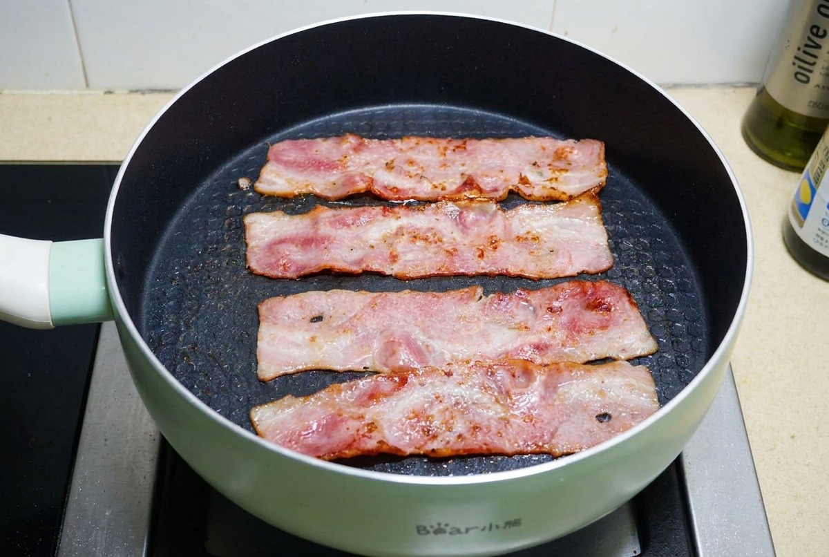 Tiras de tocino friéndose en una sartén antiadherente negra hasta que la grasa burbujee y la carne esté dorada y crujiente.
