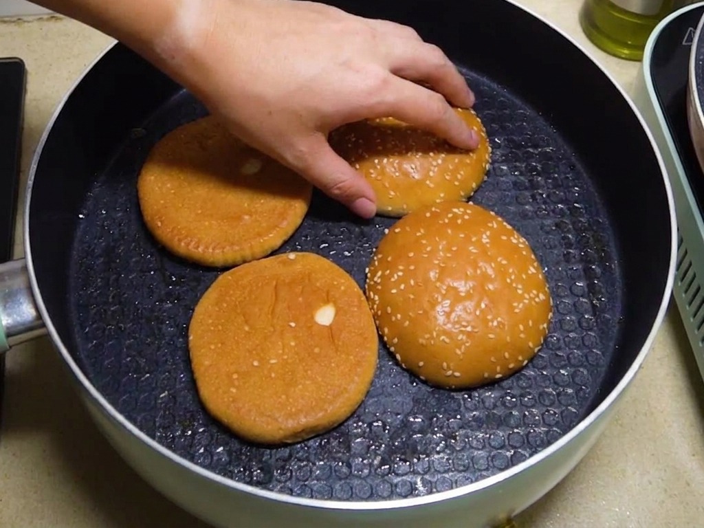 Mitades de pan de hamburguesa tostándose boca abajo en una sartén con una mano acomodándolas.