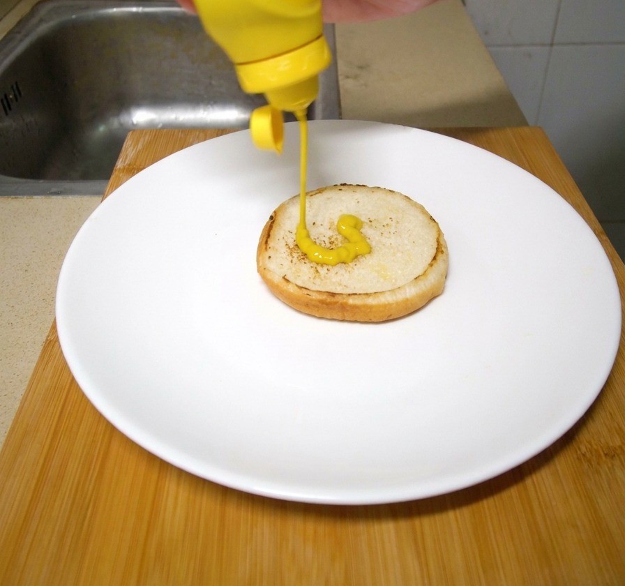 Una mano sirviendo mostaza amarilla brillante desde un envase plástico sobre un pan de hamburguesa tostado en un plato blanco.