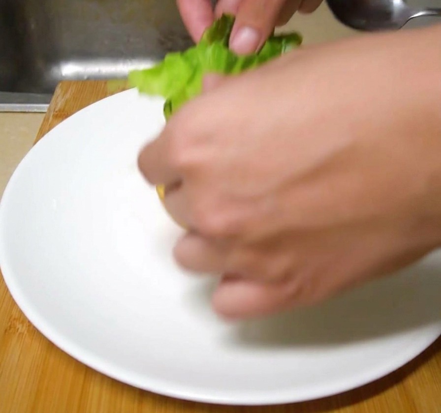 Hojas de lechuga verde fresca siendo colocadas a mano sobre un pan de hamburguesa tostado en un plato de cerámica blanco.