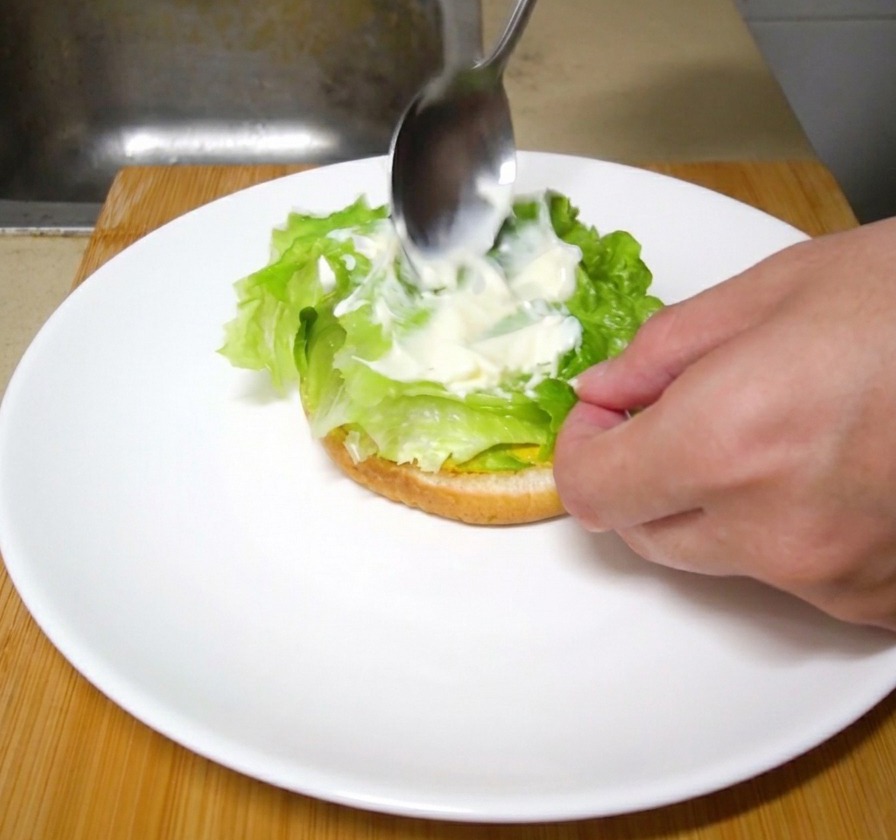 Una cuchara de metal extendiendo una porción de mayonesa blanca sobre hojas de lechuga fresca en una hamburguesa en proceso.