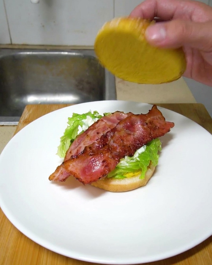 Una mano colocando la tapa de pan superior sobre una hamburguesa armada con lechuga y tiras de tocino crujiente.