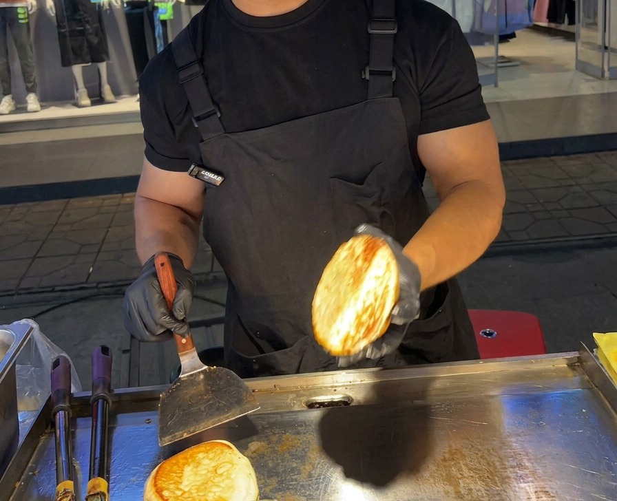 Una persona sosteniendo la mitad de un pan de hamburguesa perfectamente tostado y dorado sobre una plancha de acero inoxidable.