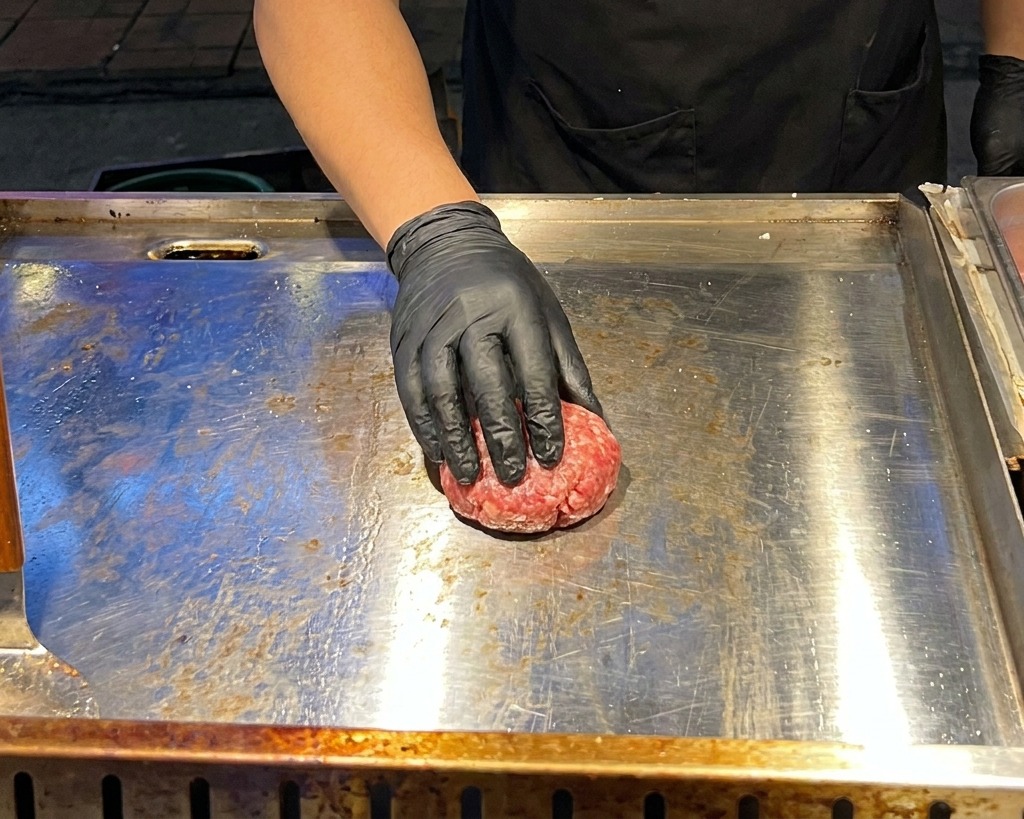 Una mano con guante negro utilizando una prensa metálica plana para aplastar una bola de carne picada sobre una plancha caliente.