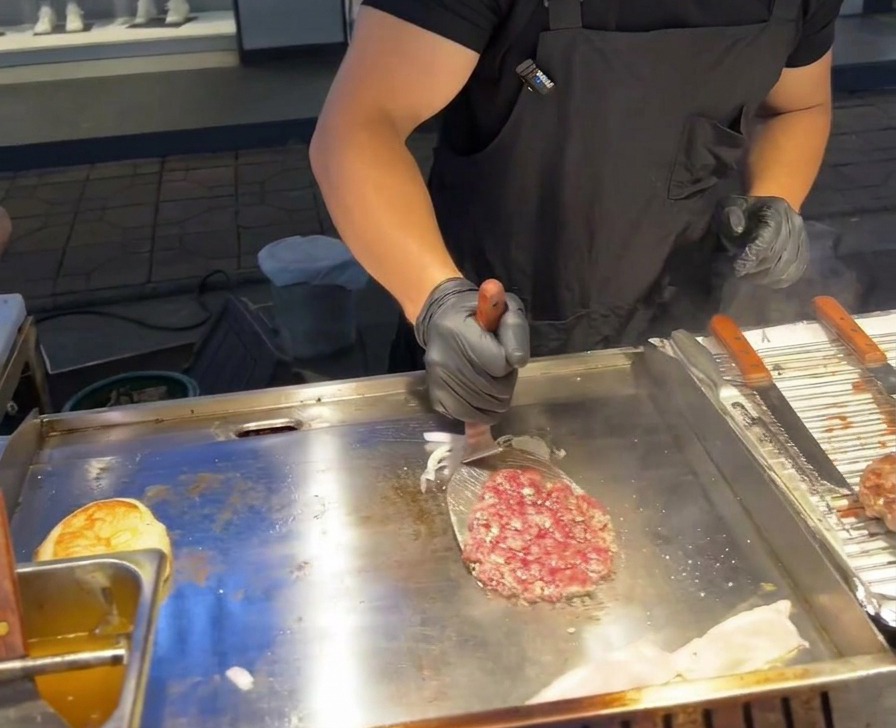 Una espátula de metal dando la vuelta a una hamburguesa de ternera sobre un montón de cebolla morada cocinada en una plancha comercial.