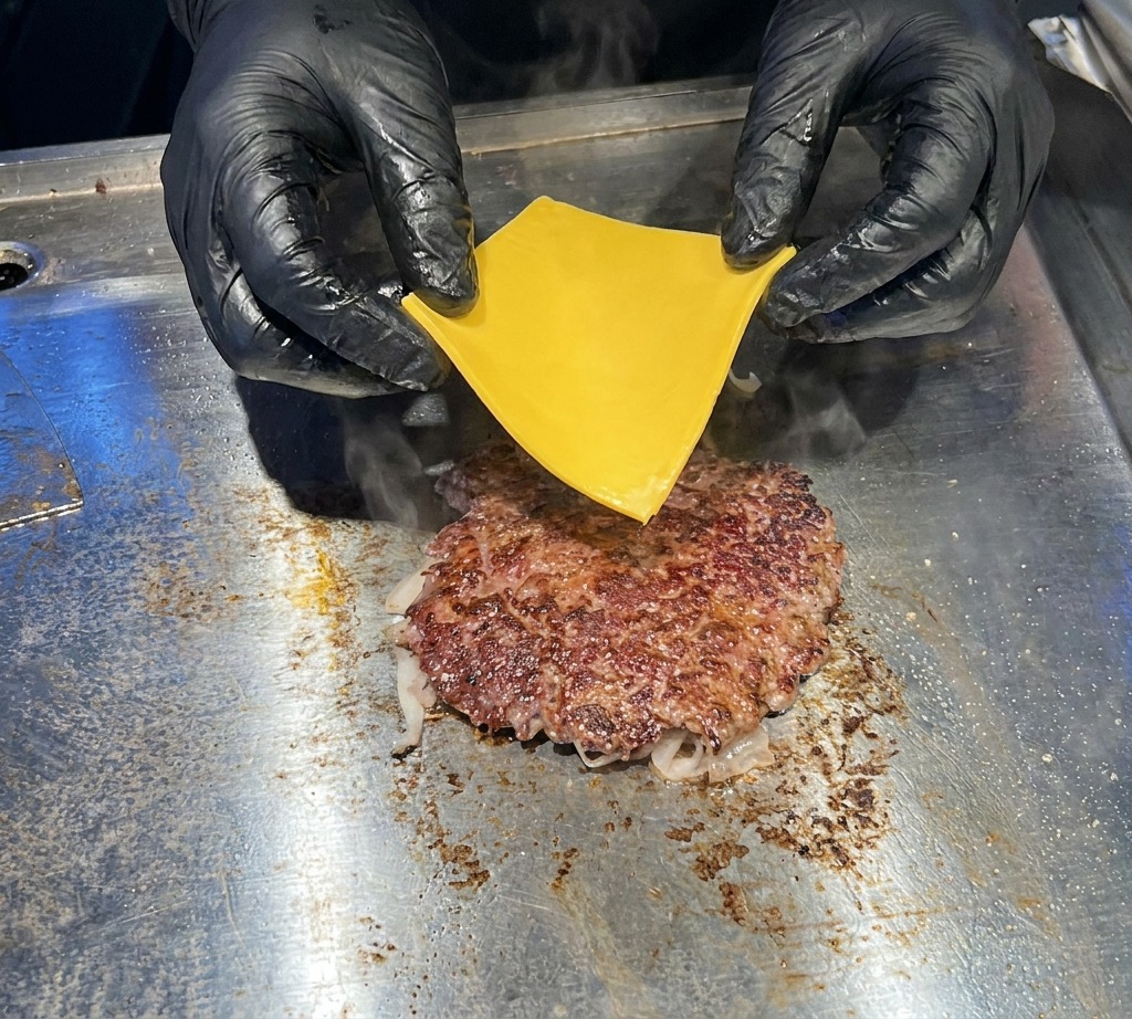 Primer plano de unas manos con guantes negros colocando con cuidado una loncha de queso americano amarillo brillante sobre una hamburguesa de carne caliente en una plancha plana.