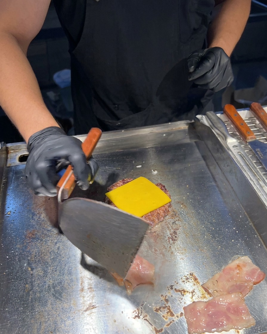 Una espátula de metal dando la vuelta a unas tiras de bacon crujiente en una plancha junto a una hamburguesa con queso amarillo fundido.