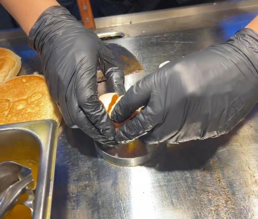 Manos con guantes negros cascando un huevo fresco dentro de un molde circular metálico plateado sobre una superficie de cocción de acero inoxidable caliente.