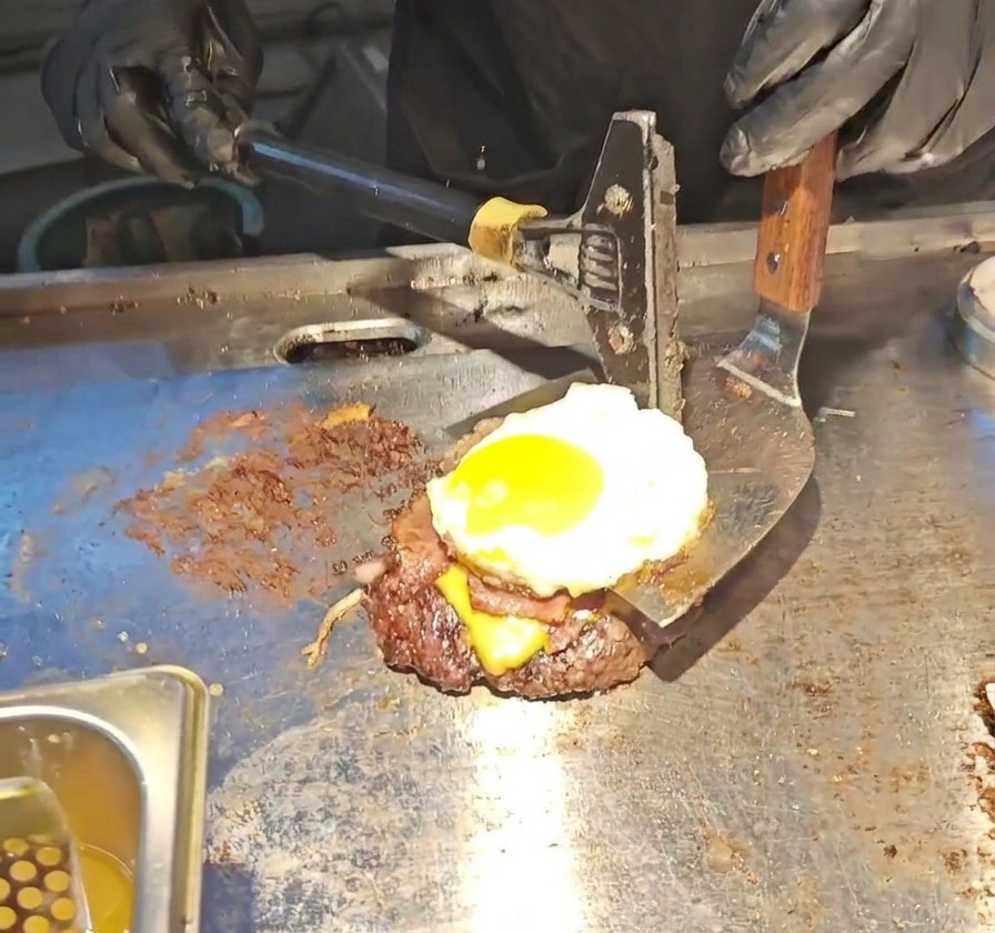 Una espátula de metal ancha levantando una torre de hamburguesa alta que consiste en carne, queso fundido, bacon y un huevo frito con yema líquida.