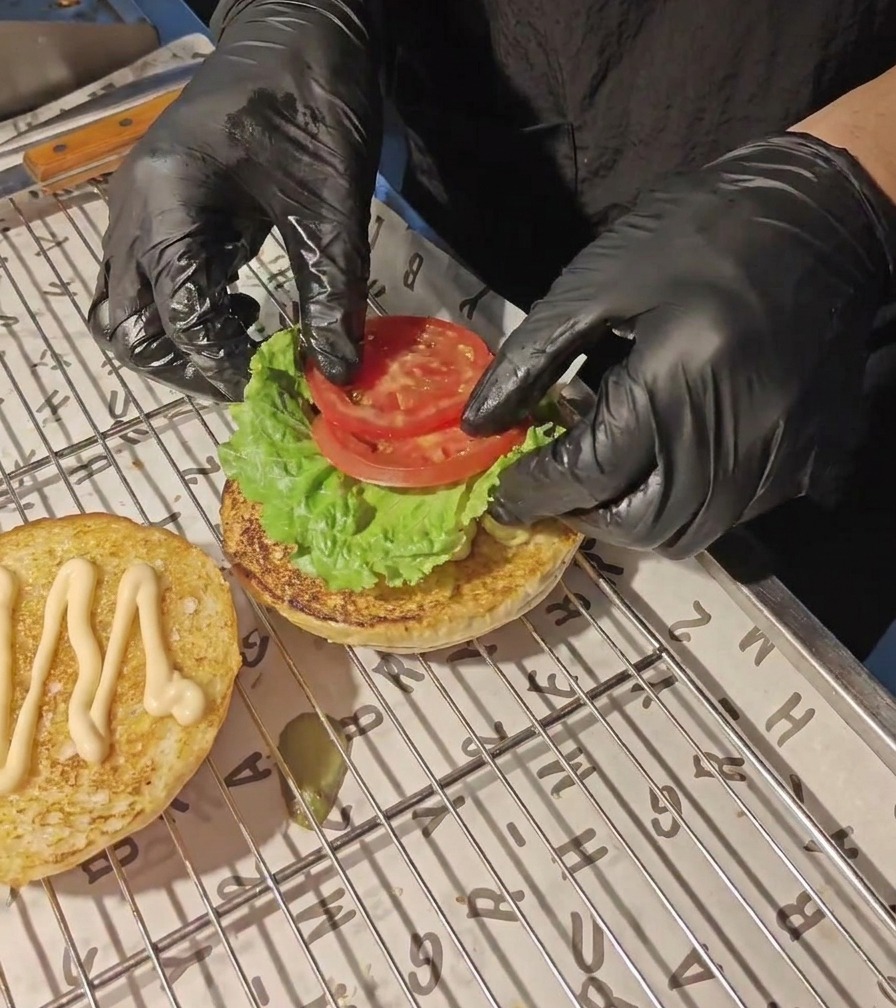 Manos con guantes negros colocando rodajas de tomate fresco sobre una hoja de lechuga verde en un pan de hamburguesa tostado.