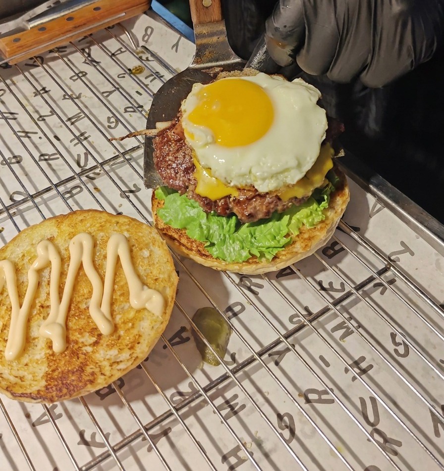 Una espátula de metal transfiriendo una hamburguesa de ternera con queso fundido y un huevo frito sobre la capa de lechuga y tomate.