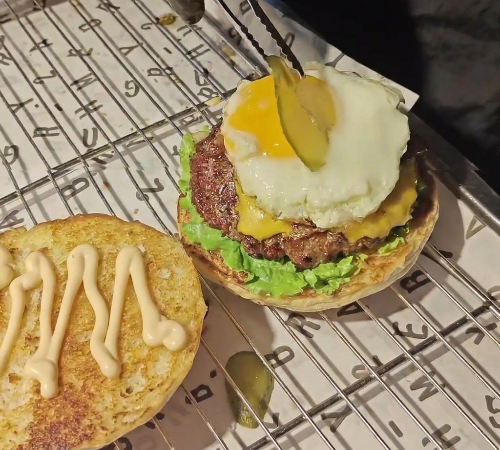 Unas pinzas colocando una rodaja fina de pepinillo sobre una yema de huevo de color amarillo brillante en la parte superior de una hamburguesa armada.
