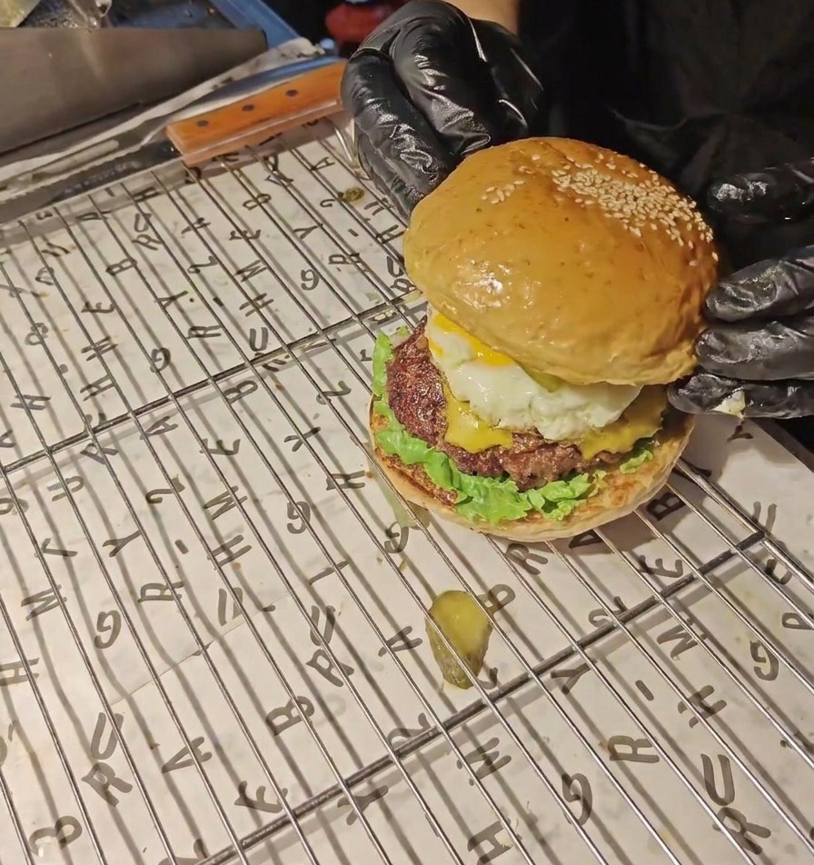 Manos con guantes negros colocando un pan dorado con semillas de sésamo en la parte superior de una hamburguesa completa de huevo y bacon.