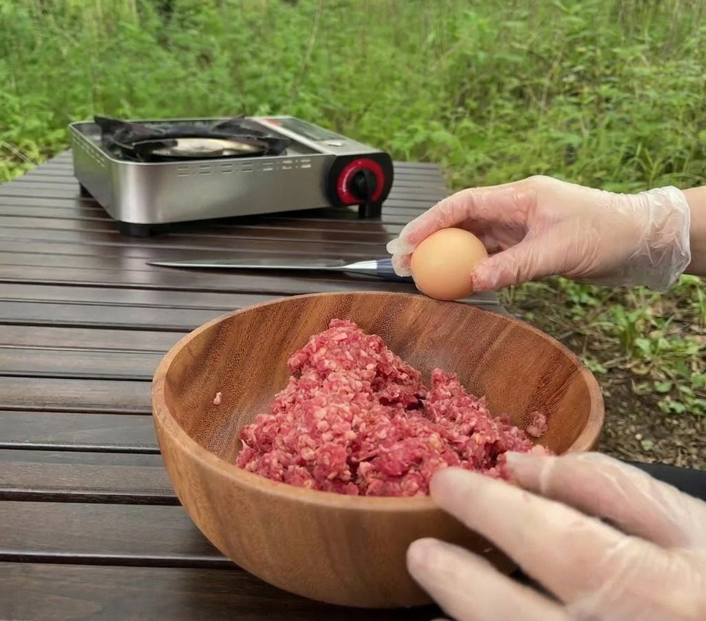 Una persona con guantes transparentes sostiene un huevo sobre un cuenco de madera lleno de carne molida cruda en una mesa al aire libre.
