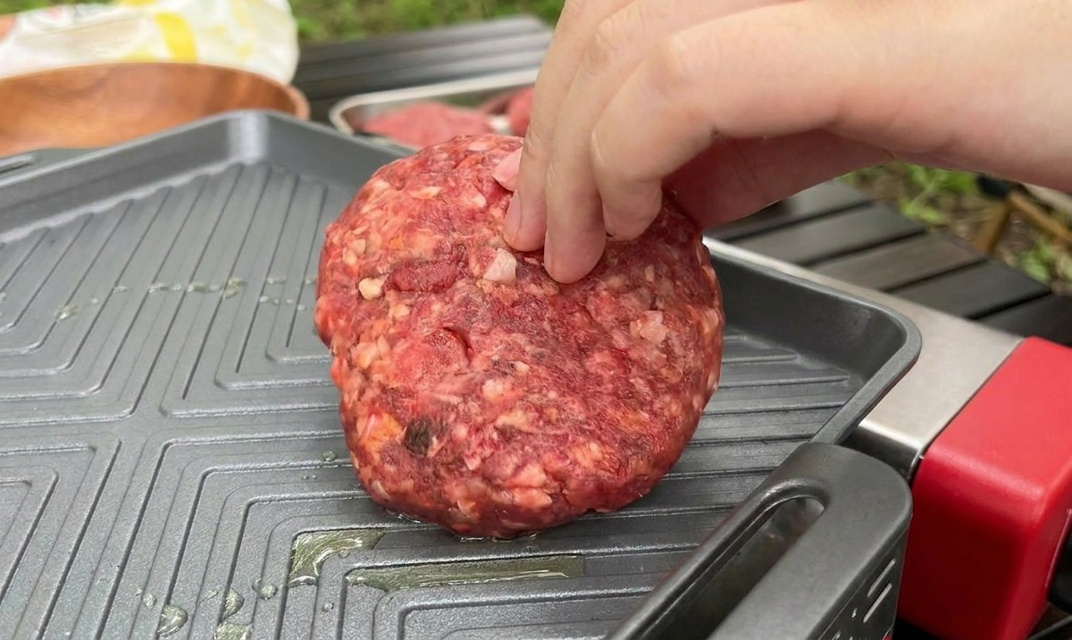 Una mano coloca una hamburguesa cruda y gruesa sobre una plancha cuadrada acanalada y caliente al aire libre.
