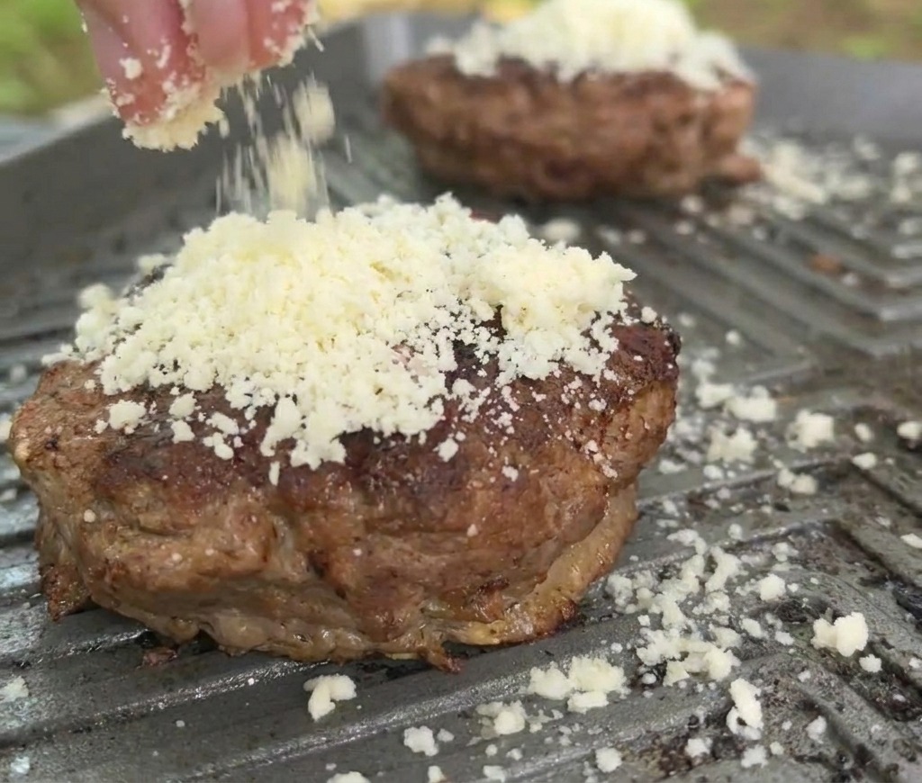 Un primer plano de queso blanco rallado cayendo sobre una carne sellada en la plancha caliente.