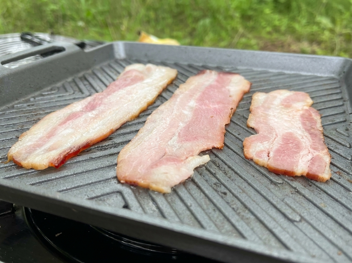 Tres largas tiras de tocino friéndose en una plancha acanalada gris, mostrando la grasa derritiéndose.