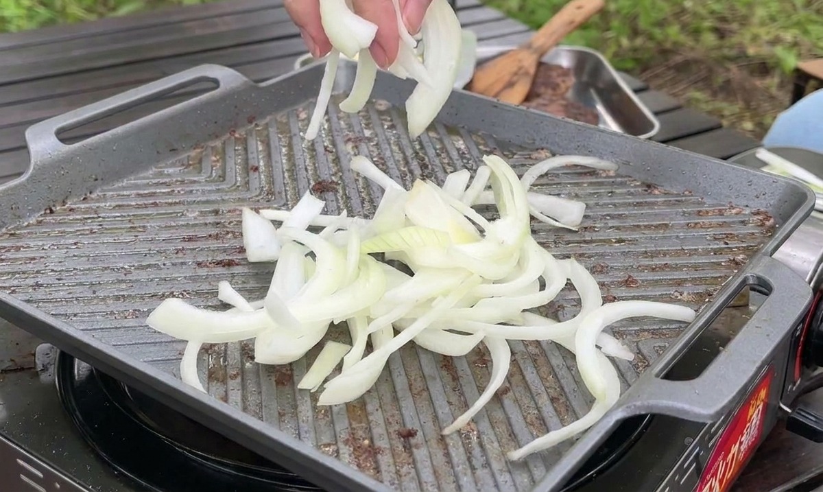 Cebollas blancas en rodajas finas siendo colocadas en una plancha caliente para saltearse junto a la carne.