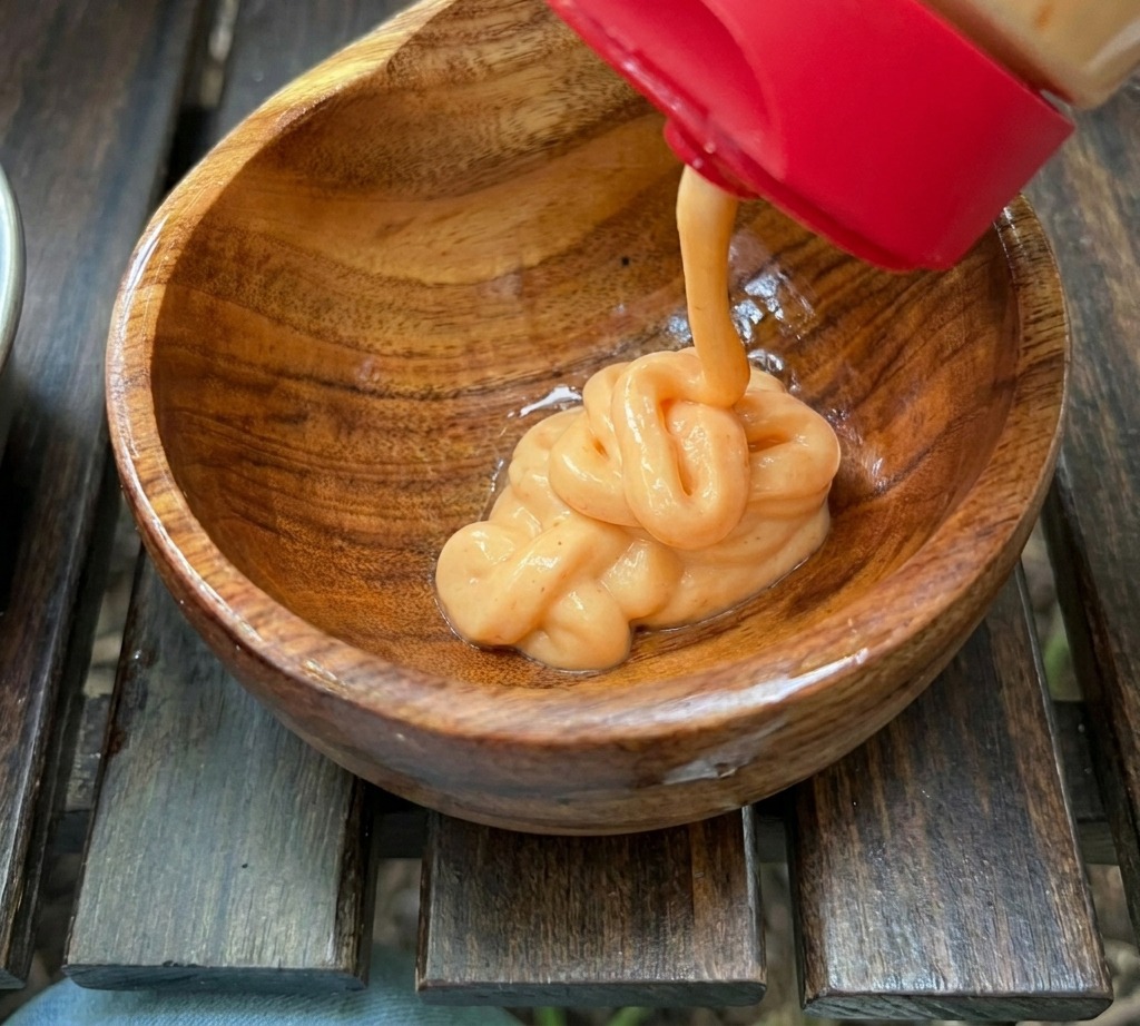 Salsa cremosa de color naranja siendo exprimida desde una botella en un pequeño cuenco de madera con otros ingredientes.