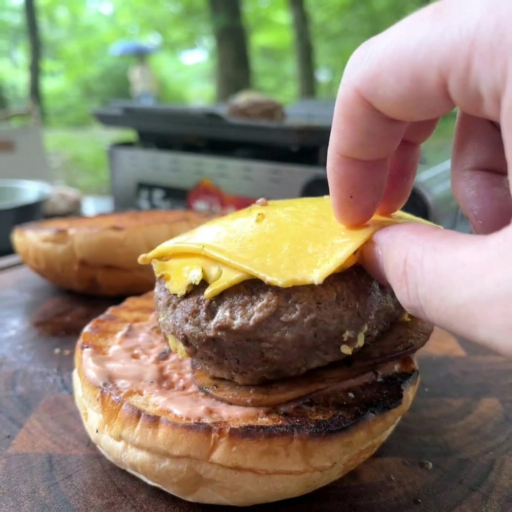Una mano coloca una carne cocida con queso amarillo fundido sobre un pan tostado untado con salsa rosa.