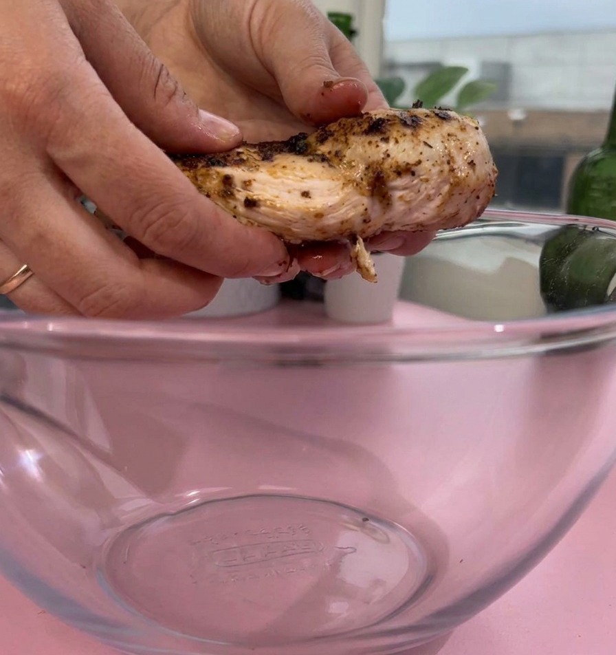 Un par de manos colocando un trozo de pechuga de pollo cocida y sazonada en un tazón de vidrio transparente sobre una superficie rosa.