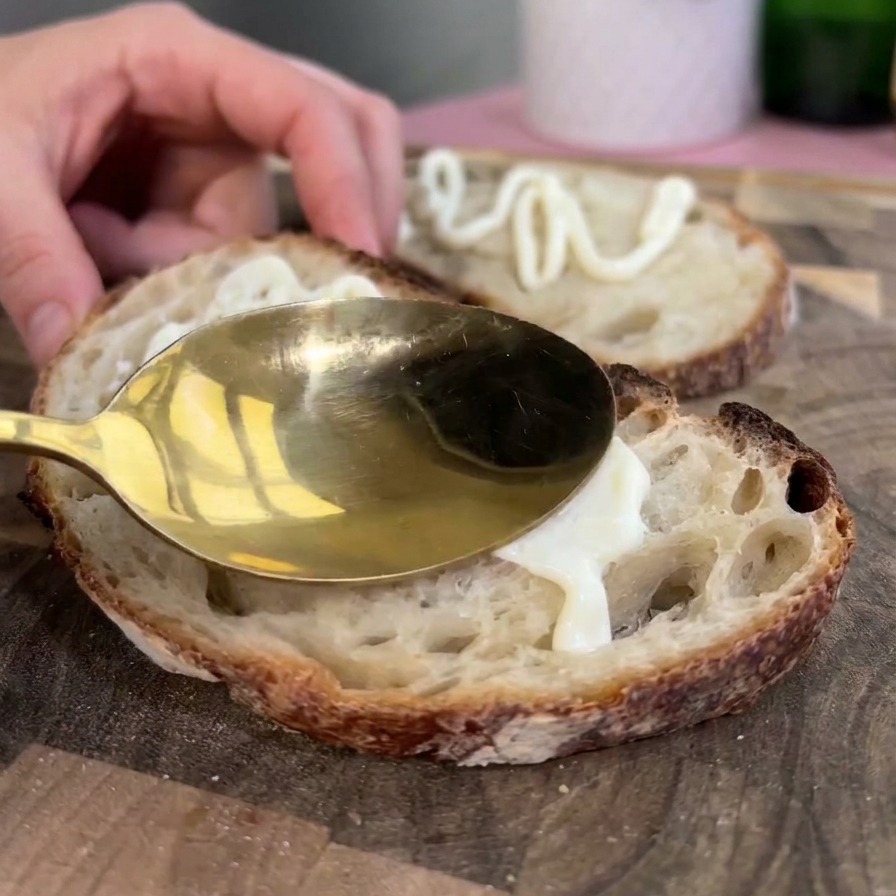 Una cuchara dorada untando una capa gruesa de mayonesa blanca sobre una rebanada tostada de pan rústico de masa madre.