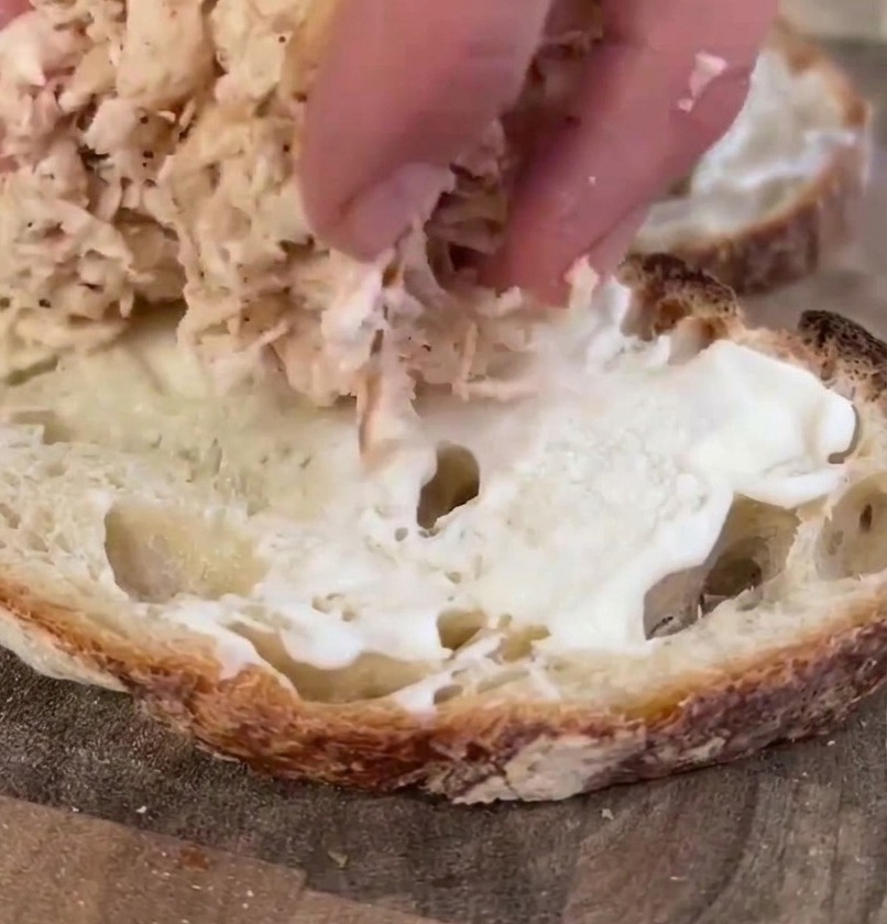 La mano de una persona colocando una porción abundante de pollo cocido y desmechado sobre una rebanada de pan de masa madre preparada con mayonesa.